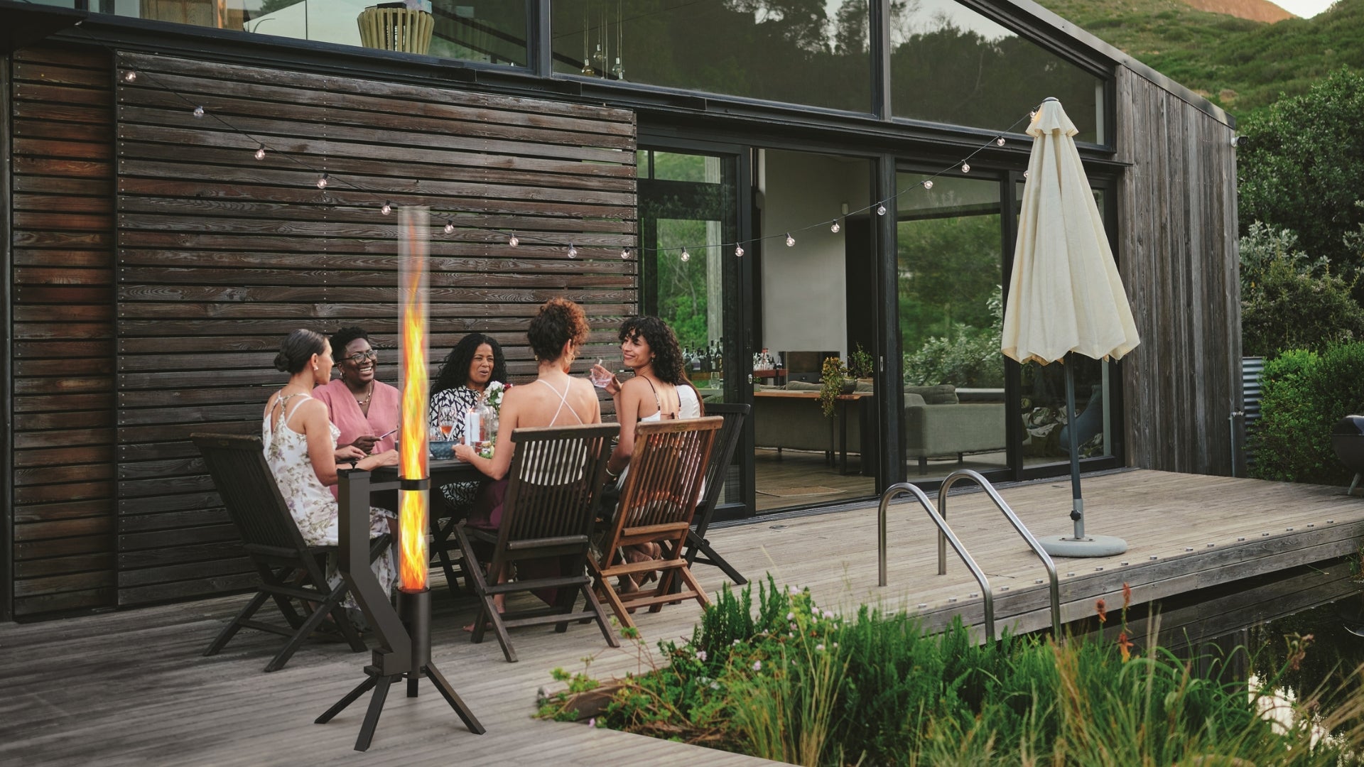 Gruppe von Freunden sitzt abends auf einer Terrasse um ein brennendes Gardenflare Feuerrohr und genießt eine gemütliche Atmosphäre