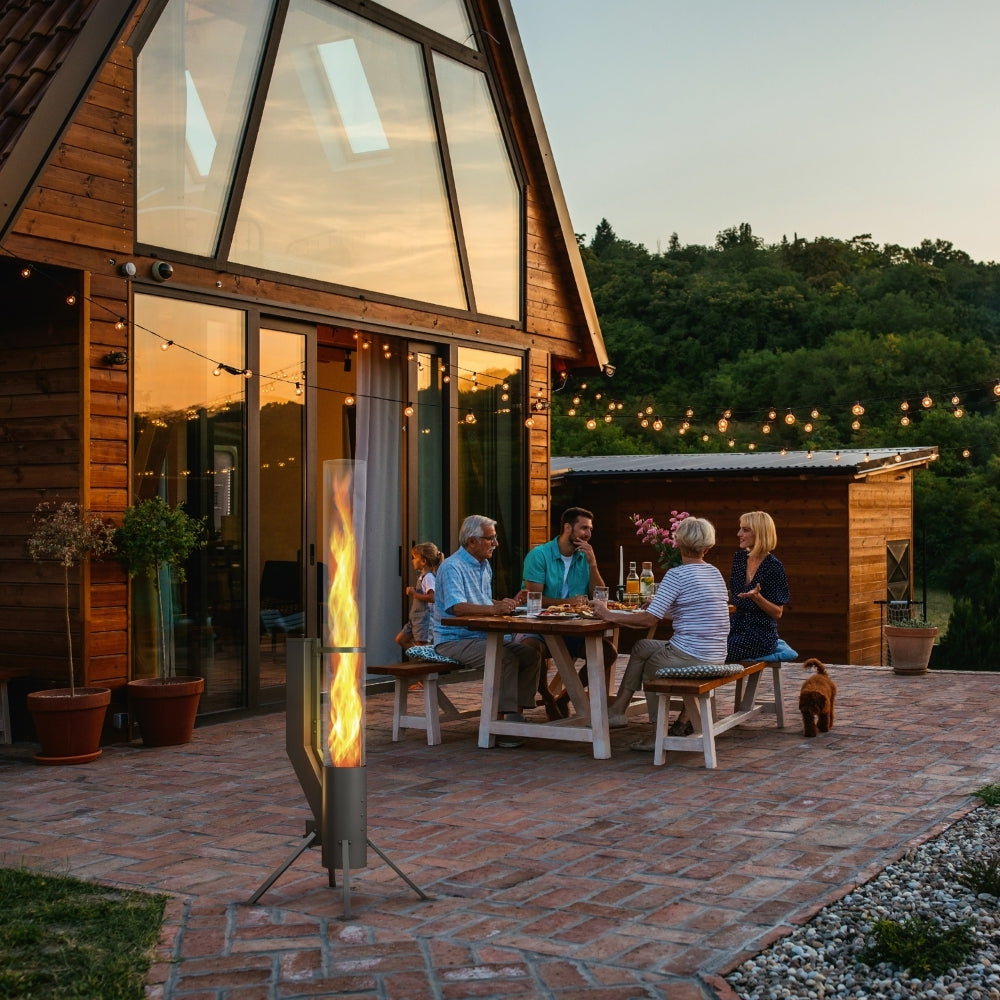 Schwarzes Gardenflare XXL Feuerrohr im Einsatz auf einer Terrasse bei Sonnenuntergang, neben einem gedeckten Gartentisch.