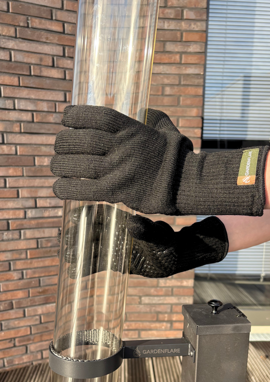 Person mit schwarzen Hitzeschutzhandschuhen von Gardenflare hält ein Glasrohr eines Feuerrohrs fest, im Hintergrund eine Hauswand aus Backstein.