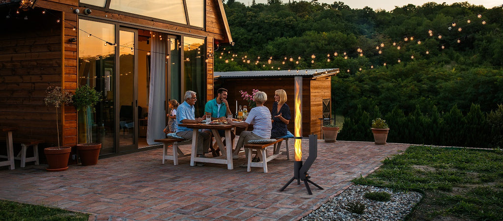Abendstimmung auf einer Terrasse mit Familie am Esstisch, im Hintergrund ein beleuchtetes Feuerrohr vor einem Holzhaus.