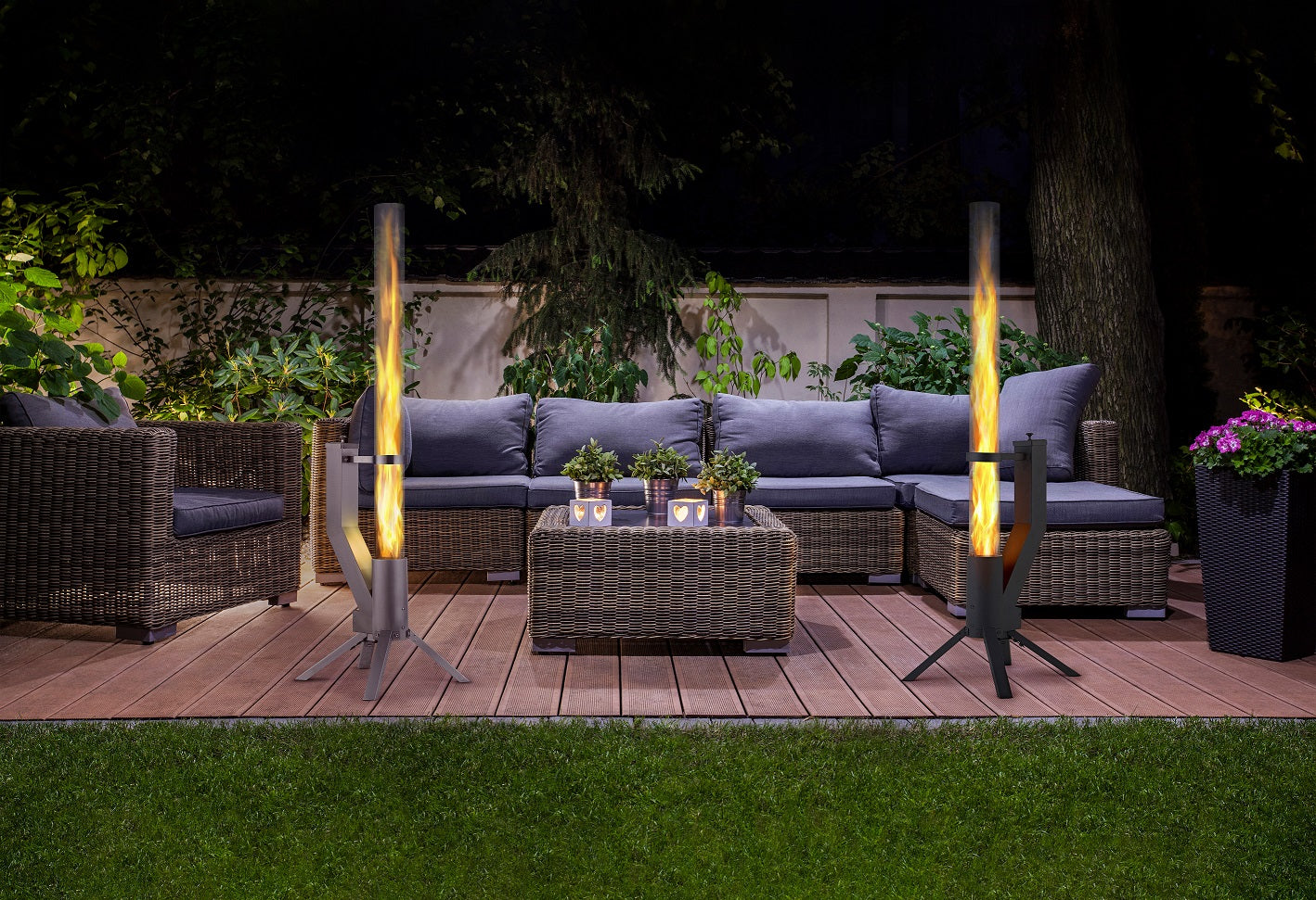 Gemütliche Terrasse bei Nacht mit Rattan-Sitzgarnitur, zwei beleuchteten Feuerrohren und dekorativen Pflanzen.